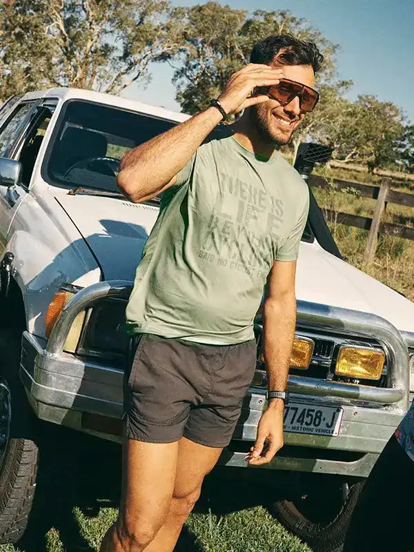 Man in olive cycling tee and shorts by vintage truck, sunny outdoor adventure wear