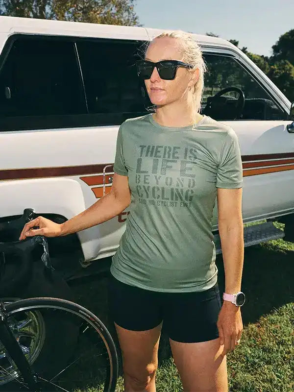 Woman in green cycling shirt with 'There Is Life Beyond Cycling' text, standing beside a bike and van