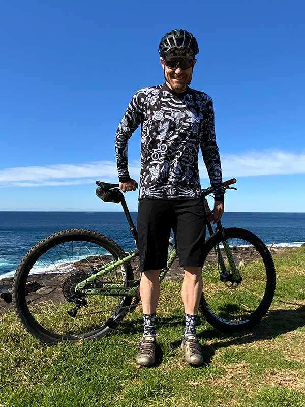 Man in patterned cycling jersey and shorts standing with mountain bike by ocean, sunny day