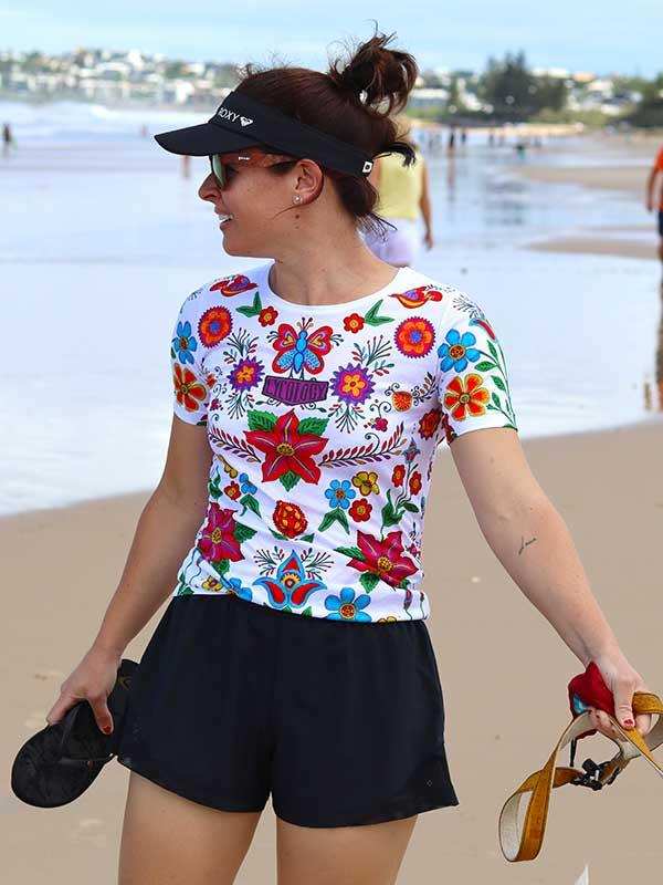 Woman wearing colorful floral cycling jersey and black shorts on beach, holding sandals