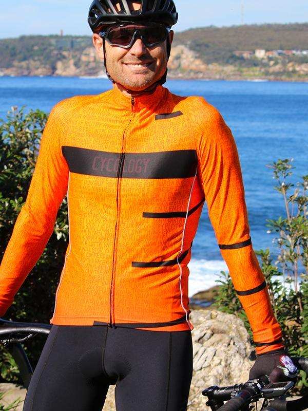 Man in orange long-sleeve cycling jersey with black accents, helmet, and sunglasses, coastal backdrop