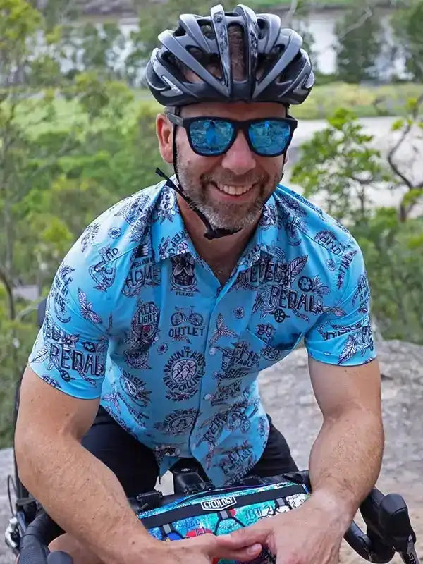 Man cycling in blue patterned jersey, helmet, and sunglasses on outdoor trail