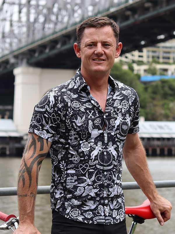 Man wearing black and white cycling-themed shirt near bridge, holding red bike seat
