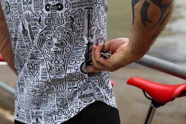 Men's cycling jersey with bold graphic print, featuring bike-themed quotes and designs, worn by cyclist near red saddle.