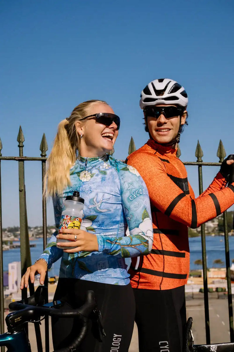 Man and woman in colorful cycling jerseys and helmets, smiling by waterfront with bikes