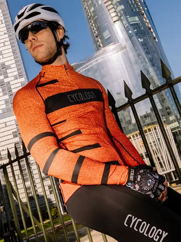 Man in orange cycling jersey and black bib shorts, wearing helmet and gloves, urban city backdrop