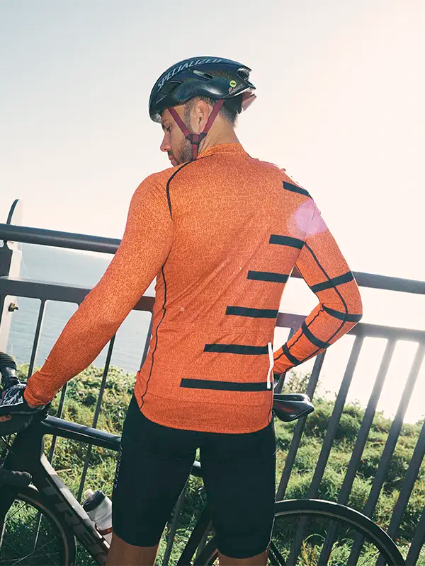 Person wearing an orange cycling jersey with black stripes, standing on a bridge with a scenic background.