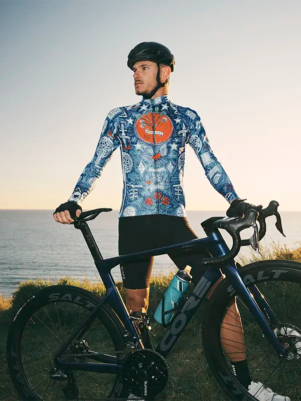 Man in a colorful cycling outfit standing next to a bicycle by the sea.