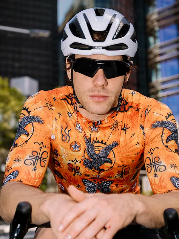 Person wearing an orange cycling jersey with a helmet and sunglasses, sitting on a bike.