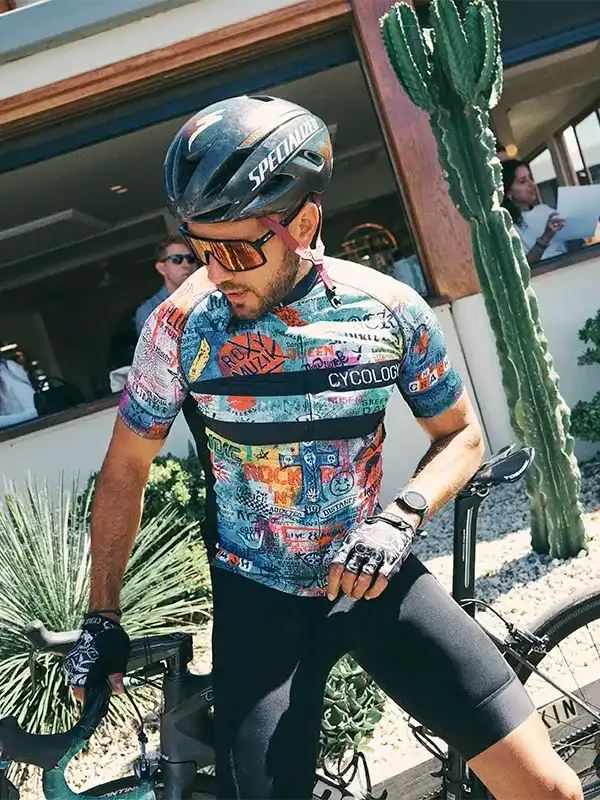 Man in colorful cycling jersey and helmet, standing with bike near cactus, wearing gloves and sunglasses