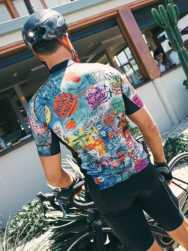 Person wearing a colorful cycling jersey with various graphics and text, standing next to a bicycle.