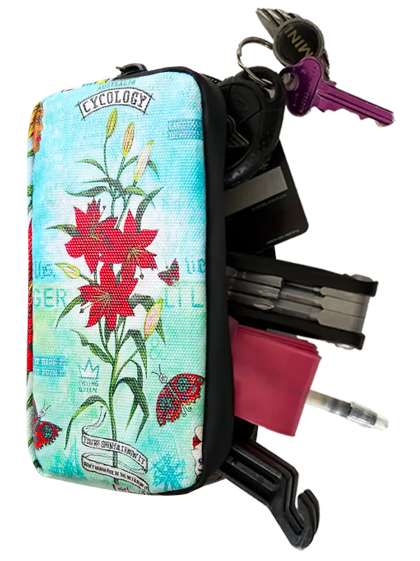 A small zippered cycling wallet with a light blue botanical print featuring red lilies, green leaves, butterflies, and decorative text. The wallet is shown open with various keys, a black wallet, a pink card holder, and a pen sticking out, set against a plain white background.