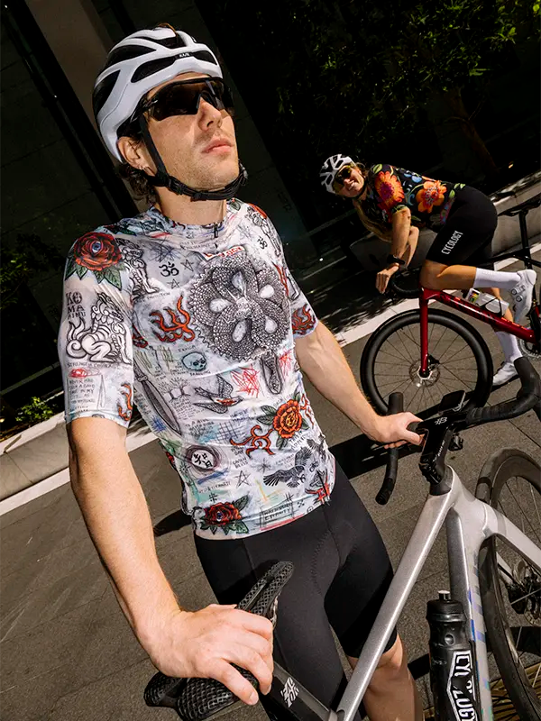Cyclist wearing a gray cycling jersey with various designs and helmet with another cyclist in the background.