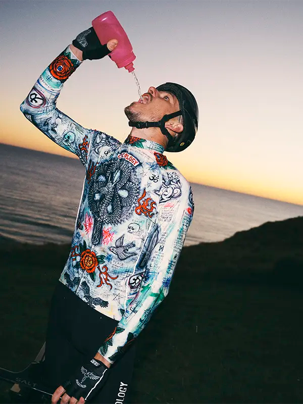 Person wearing a colorful jersey with a helmet, pouring a pink bottle against a sunset sky.