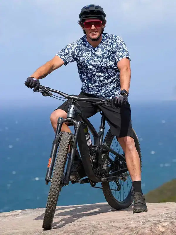 Man in floral cycling jersey riding mountain bike on cliff overlooking ocean, wearing helmet and sunglasses