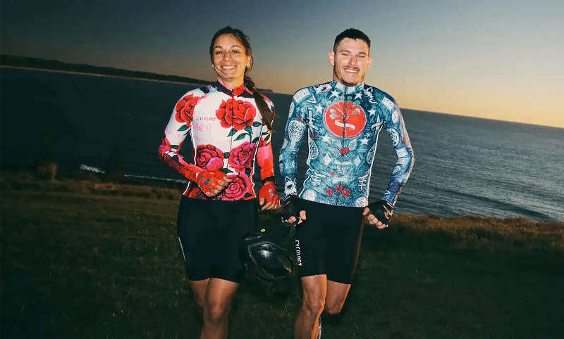 Man and woman in colorful cycling jerseys and shorts, smiling by the ocean at sunset, holding helmets.