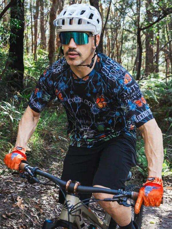 Man riding mountain bike in forest wearing floral cycling jersey, helmet, and orange gloves