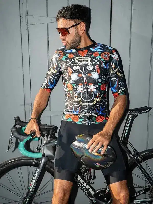 Man in colorful cycling jersey and shorts holding helmet beside road bike