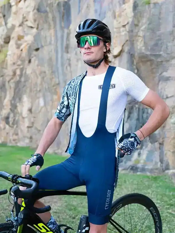 Man in cycling bib shorts and jersey with helmet, sunglasses, gloves, standing beside road bike outdoors