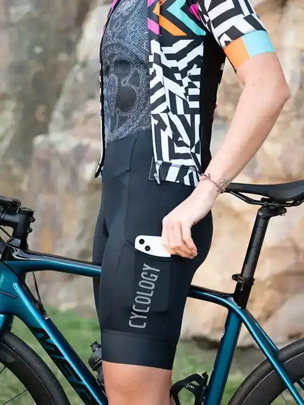Cyclist wearing patterned jersey and black cycling shorts with phone pocket, standing beside road bike