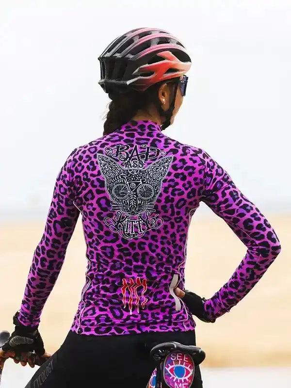 Woman wearing purple leopard print cycling jersey with 'Bad Ass Kitty' graphic, helmet, and gloves on bike