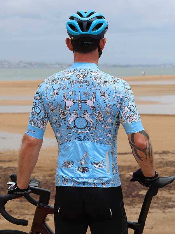 Men's light blue cycling jersey with tattoo-style graphics, worn on beach with bike